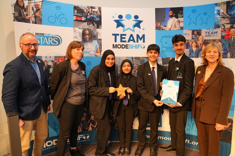 A group of seven people, including five adults and two students, posing in front of a backdrop featuring the "Team ModeShift Stars" logo. The students, wearing black school uniforms, are holding a certificate and a star-shaped award. The adults, including two women wearing business attire and a man in a blazer, are flanking the students, all smiling. The backdrop features additional images, including people, transportation, and logos.