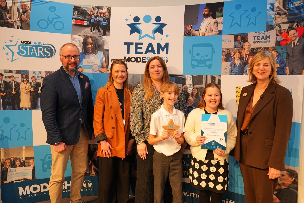 A group of six people, consisting of four adults and two children, pose for a photograph in front of a blue backdrop with promotional graphics for the "Team Modeshift STARS" program. The children, a girl and a boy, hold up awards, and the adults give thumbs up. The backdrop features images of buses, bikes, and other transportation-related elements, along with the "Team Modeshift" logo and the program's name.