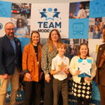 A group of six people, consisting of four adults and two children, pose for a photograph in front of a blue backdrop with promotional graphics for the "Team Modeshift STARS" program. The children, a girl and a boy, hold up awards, and the adults give thumbs up. The backdrop features images of buses, bikes, and other transportation-related elements, along with the "Team Modeshift" logo and the program's name.