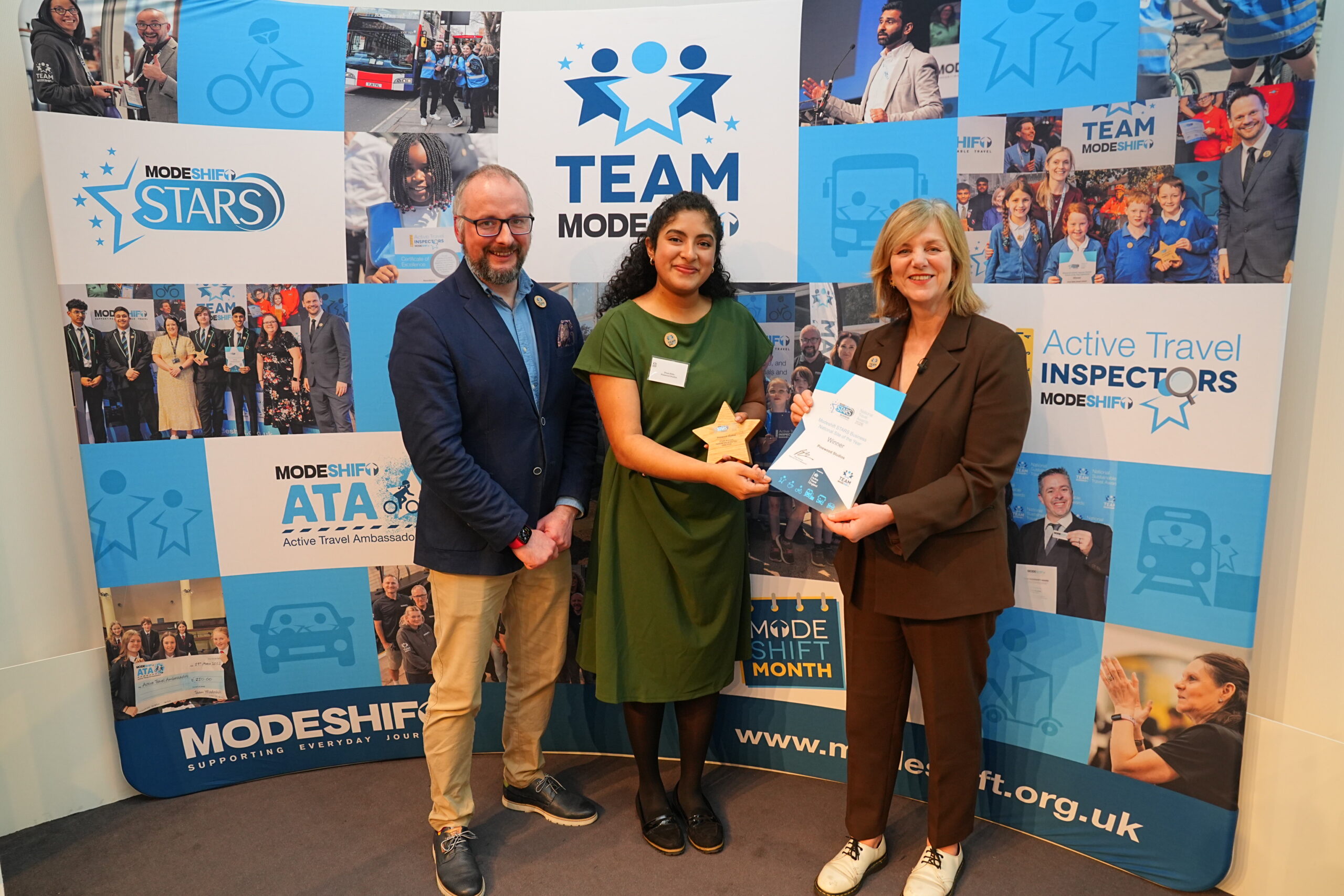 Three people posing for a photo in front of a promotional banner. A woman in a green dress and black shoes holds a wooden star, a man in a navy blazer and khakis stands to the left, and a woman in a brown suit is on the right, holding a certificate. The banner behind them shows the "Modeshift STARS" and "Team Modeshift" logos, along with images of people and transportation methods like bikes, buses, and trains. The certificate is a "Modeshift STARS Business National Site of the Year" award.