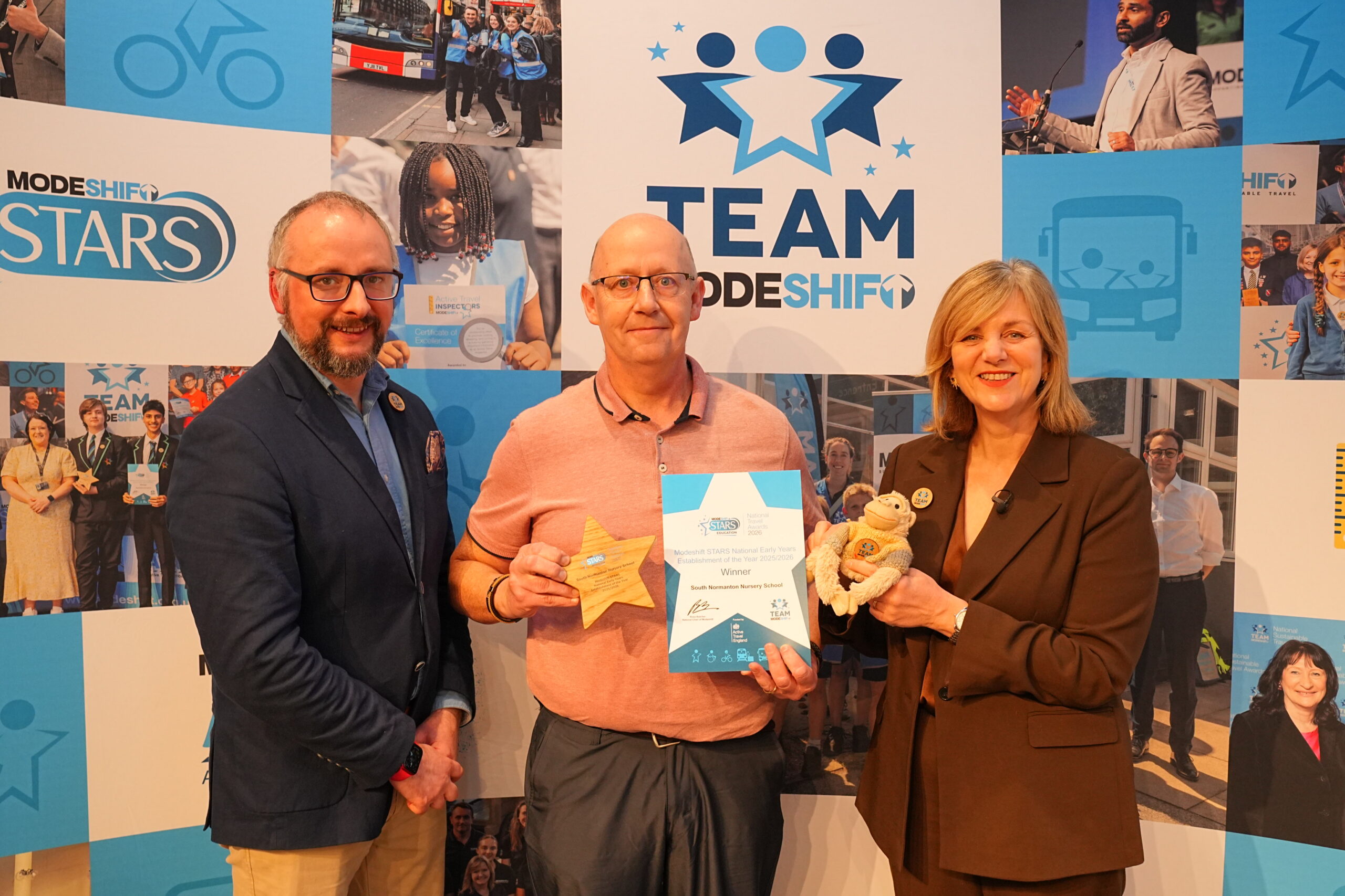 South Normanton Nursery School - Modeshift STARS Early Years Establishment of the Year 2025/26. Three people, two men and a woman, stand in front of a blue banner, smiling and holding awards. The central man holds a wooden star-shaped award and a certificate that reads "Modeshift STARS National Early Years Establishment of the Year 2025/2026 Winner South Normanton Nursery School." The woman beside him holds a monkey plush toy and wears a brown suit. The man to her left, wears a blue suit jacket and glasses. The banner behind them has "TEAM MODESHIFT" written on it along with the "STARS" logo.