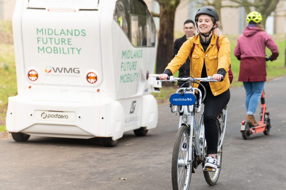 Person wearing a yellow jacket and helmet rides a Nextbike bicycle on a paved path near an autonomous vehicle labeled Midlands Future Mobility.