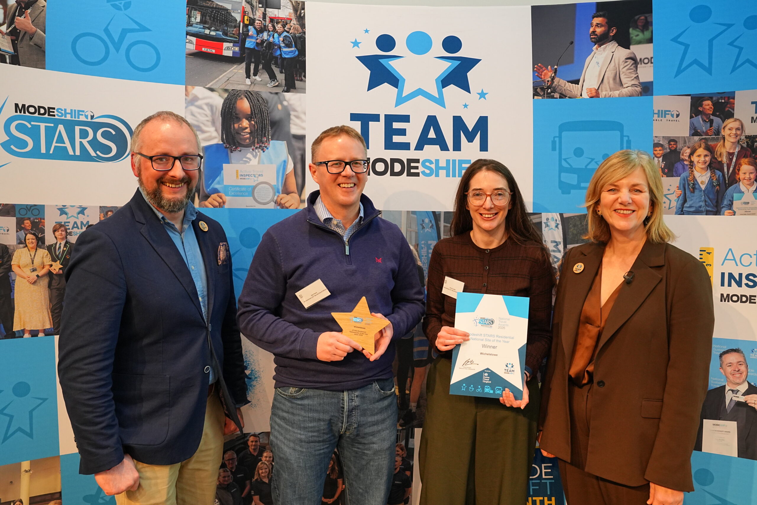 Four people smiling and posing for a photo at an awards ceremony, standing in front of a blue background with the text "TEAM MODESHIFT STARS" and images related to travel. A man on the left and a woman on the right are wearing suit jackets, and two middle people are dressed more casually. The middle two are holding a star-shaped award and a certificate with "National Site of the Year" printed on it.