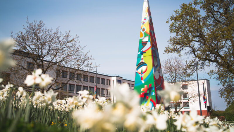 Colorful tall sculpture with abstract designs stands in front of a modern building surrounded by blooming white flowers and trees under a clear sky.