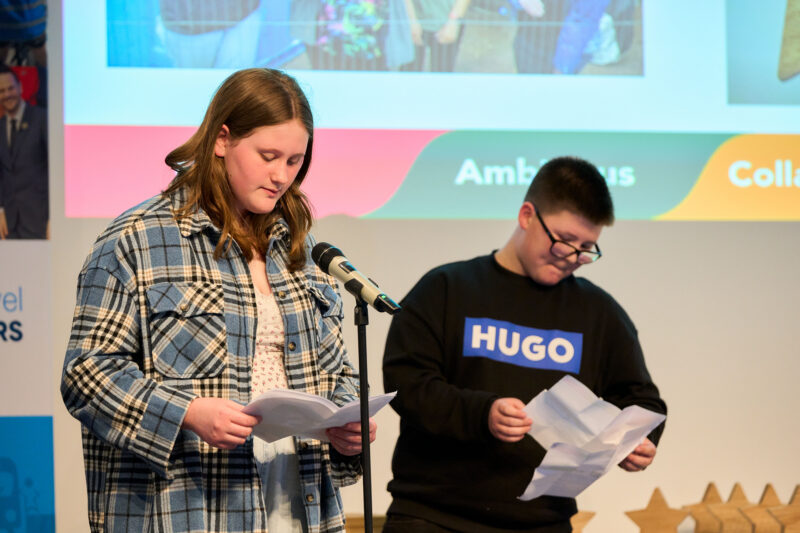 Two young people stand at a podium, each holding and reading from a piece of paper, with a projector screen behind them showing abstract shapes and text. Aurora Hedgeway School - Modeshift STARS National SEND School of the Year 2025/26
