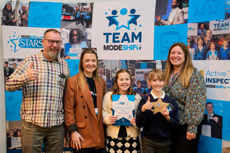 Outwoods Primary - Modeshift STARS National Primary School of the year. Four people smile for a photo in front of a backdrop with "MODESHIFT STARS" and "TEAM MODESHIFT" logos. Two children in the center hold certificates and a star-shaped award, with adults on either side. The backdrop features various images related to cycling and transportation. 