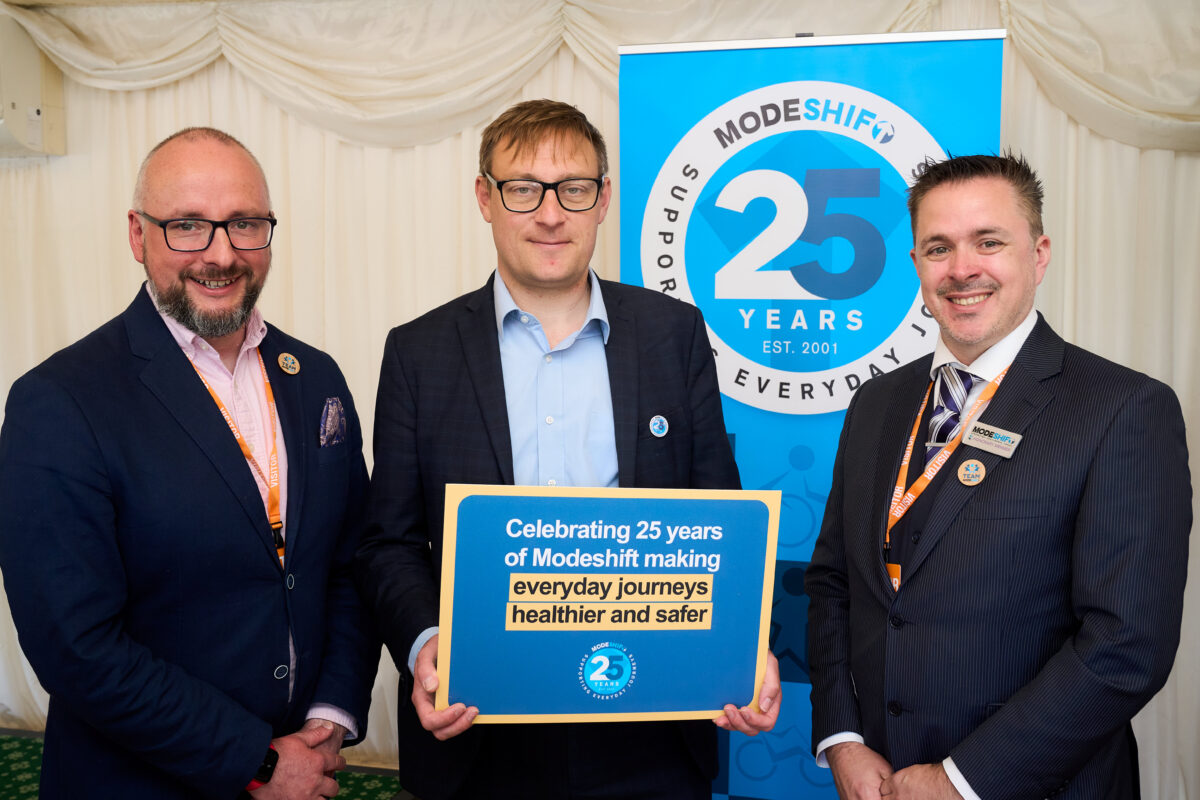 Three men in suits stand in front of a blue banner celebrating "Modeshift 25 Years Est. 2001" and holding a blue sign that reads "Celebrating 25 years of Modeshift making everyday journeys healthier and safer."