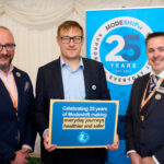 Three men in suits stand in front of a blue banner celebrating "Modeshift 25 Years Est. 2001" and holding a blue sign that reads "Celebrating 25 years of Modeshift making everyday journeys healthier and safer."