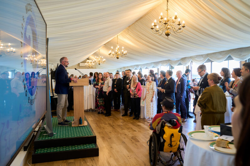 A man stands at a wooden podium on a raised platform, speaking to an audience gathered under a draped ceiling with chandeliers. To the left, a large screen displays a swirling logo with text, reflecting the audience. The audience members are dressed in formal and semi-formal attire, with some standing and others seated. One person in the foreground is in a wheelchair.