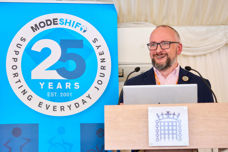 Man in a suit speaking at a wooden podium with microphones and a laptop. A blue banner with "MODESHIFT" and "25" is behind him. A crest of the Houses of Parliament is on the podium.