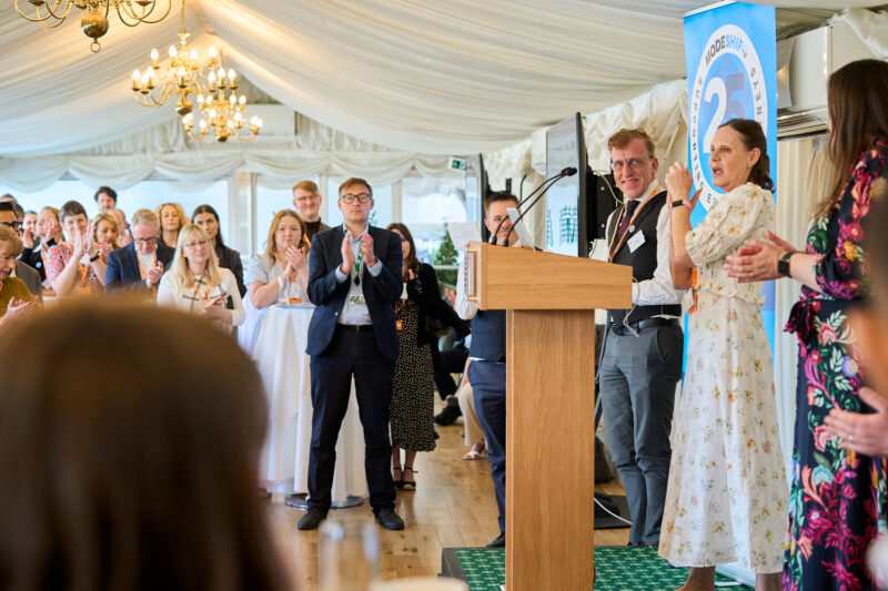 Attendees applaud at an event with a speaker at a podium and a banner in the background.