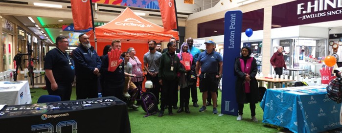 A group of people stand on green carpet in a shopping center near promotional tents and tables displaying branded materials, including one with "Stagecoach" visible.