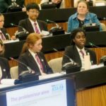 A group of students in school uniforms sit at a table with microphones, presenting in a formal setting.