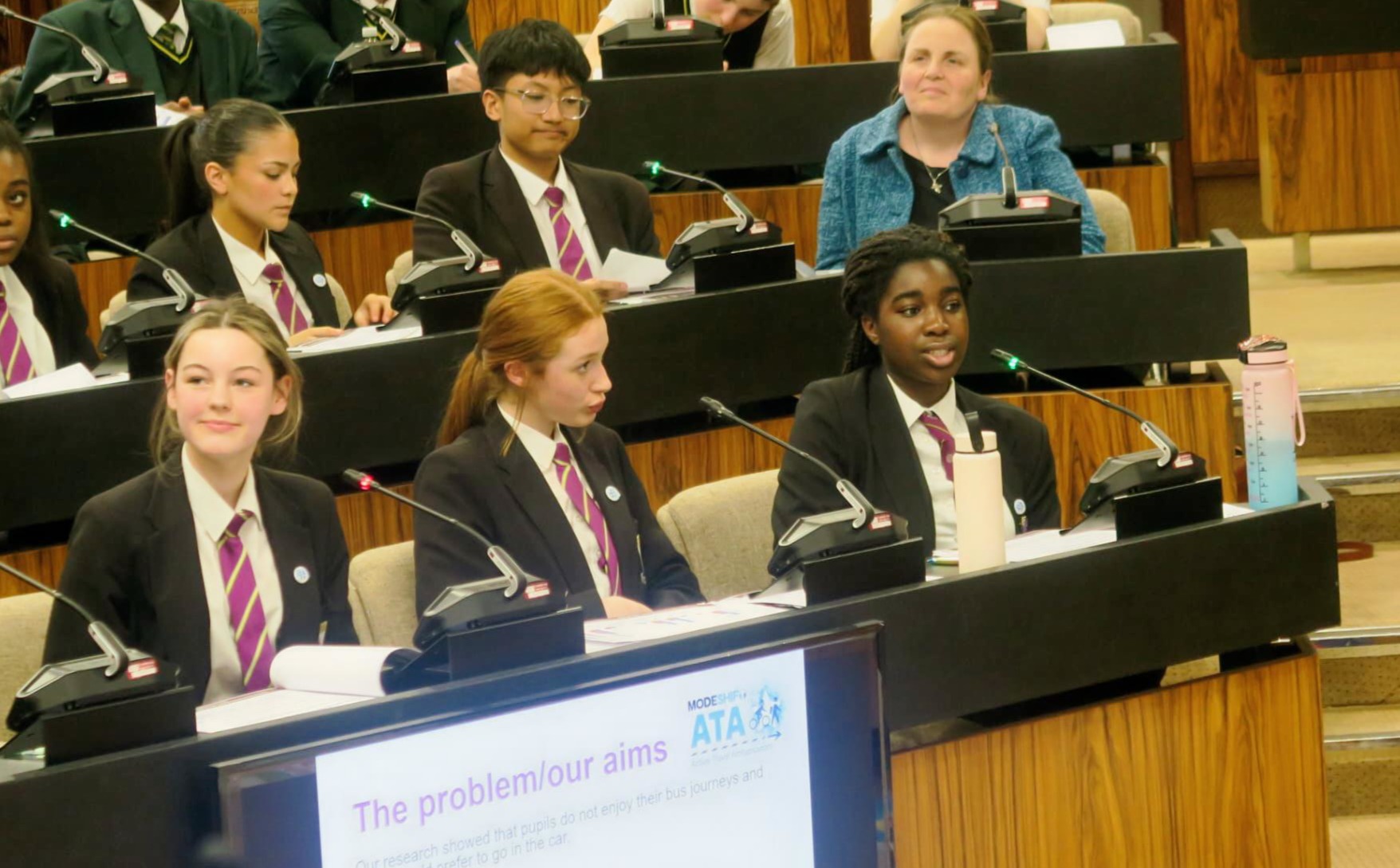 A group of students in school uniforms sit at a table with microphones, presenting in a formal setting.
