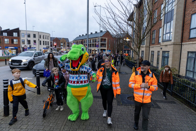 A large, friendly green alligator mascot in a festive, colorful sweater walks alongside people on a paved walkway in front of brick buildings. Telford and Wrekin – Celebrating their first STARS accredited school.