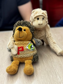 Two stuffed animals, a hedgehog wearing a sweater and a monkey, sit side-by-side on a wooden surface.