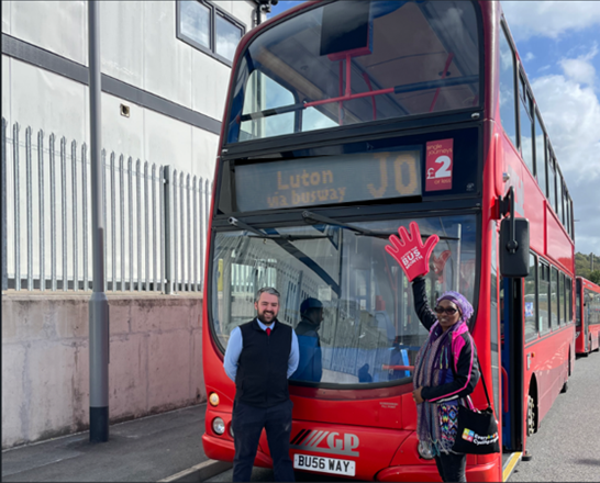 Two people stand in front of a red double-decker bus with "Luton via busway J0" displayed on the front. The person on the left, appearing to be a bus driver in uniform, smiles at the camera. The person on the right, wearing a purple headscarf and holding a red hand-shaped foam finger with "BUS" printed on it, waves.