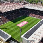 Aerial view of a football stadium filled with spectators, with a large pink and yellow banner unfurled on the green pitch.
