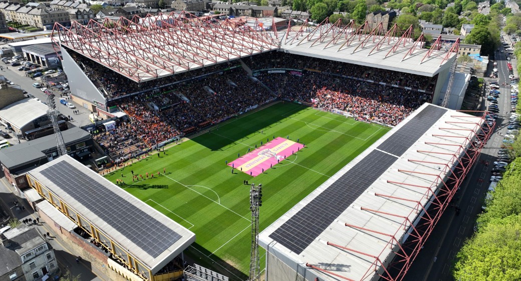 Aerial view of a football stadium filled with spectators, with a large pink and yellow banner unfurled on the green pitch.