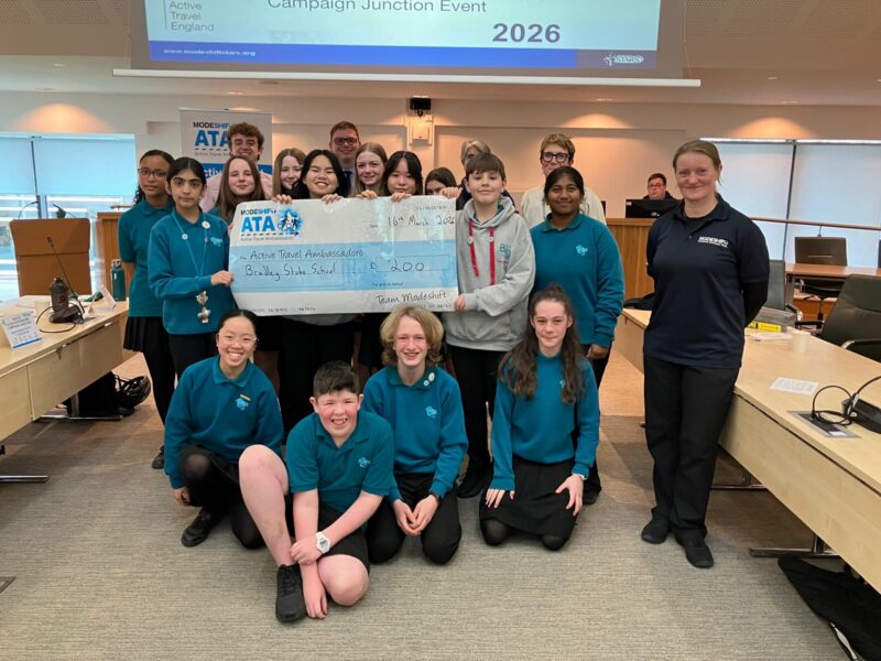 A group of young people and adults stand together, with several holding a large novelty cheque for £200 made out to "Active Travel Ambassadors, Bradley Stoke School, Team Modeshift." The event appears to be a "Campaign Junction Event" with the year 2026 displayed prominently on a screen in the background.