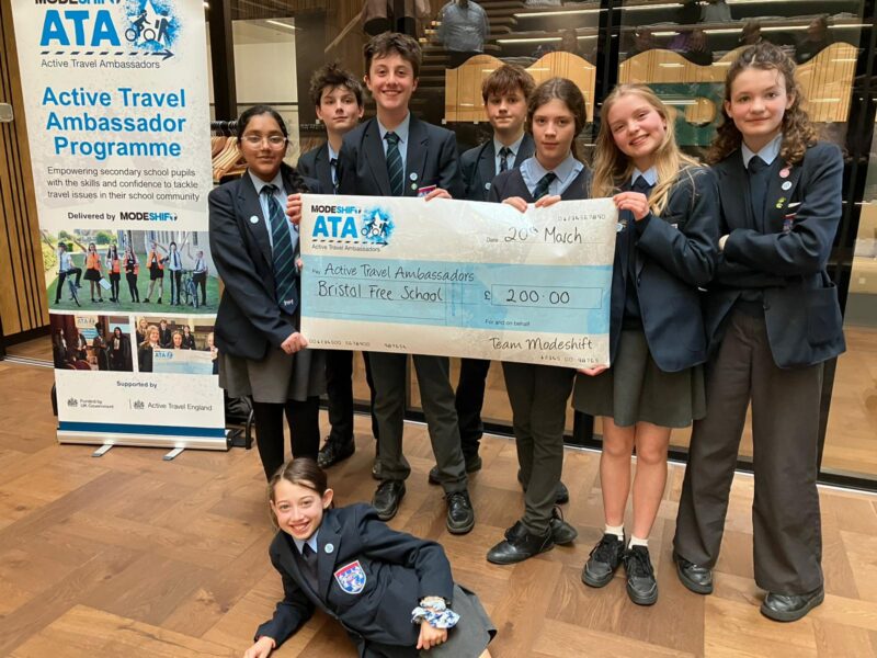 A group of students in uniform stand behind a large novelty check for £200.00, made out to "Active Travel Ambassadors Bristol Free School" and signed "Team Modeshift," with a banner in the background promoting the "Active Travel Ambassador Programme."