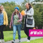 Three women walking together on a park path surrounded by green trees during National Walking Month.