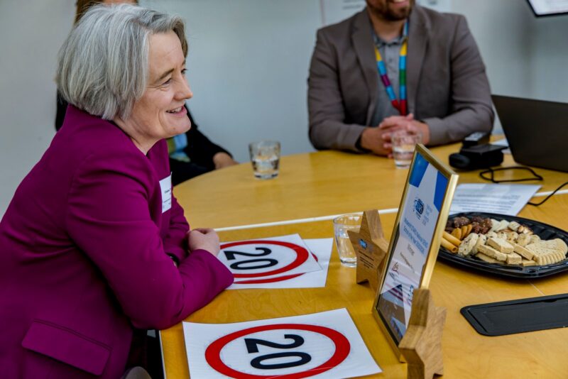 A woman in a magenta blazer smiles while leaning on a wooden table, where two "20" speed limit signs, glasses of water, a platter of cookies, and framed certificates are placed.