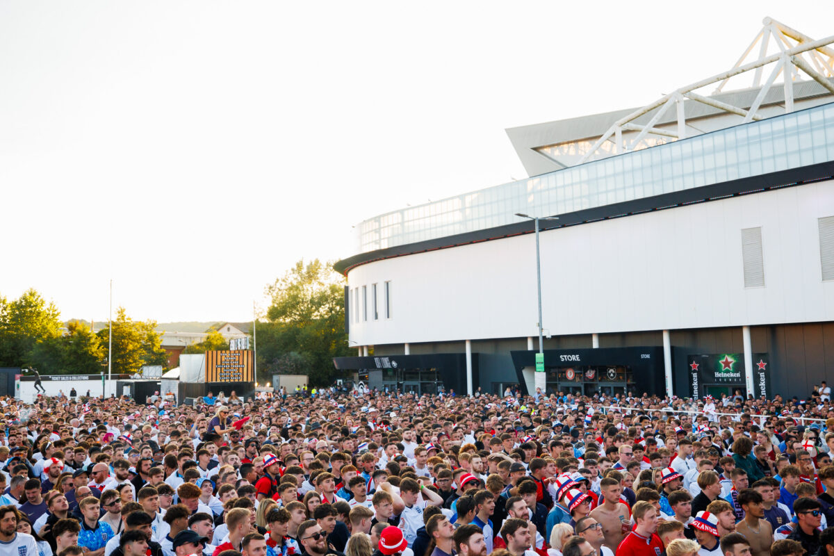 A large, diverse crowd of people, many wearing England football team colors and hats, gather outdoors in front of a modern stadium on a sunny day. Ashton Gate Stadium joins Modeshift STARS Leisure Membership