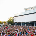 A large, diverse crowd of people, many wearing England football team colors and hats, gather outdoors in front of a modern stadium on a sunny day. Ashton Gate Stadium joins Modeshift STARS Leisure Membership