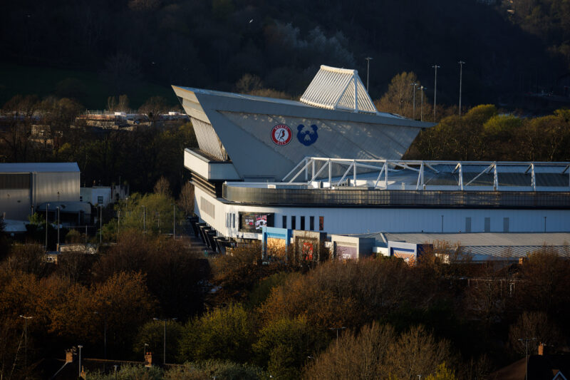 Modern sports stadium with a distinctive triangular roof and large logos on its facade, surrounded by trees and buildings under a clear sky.