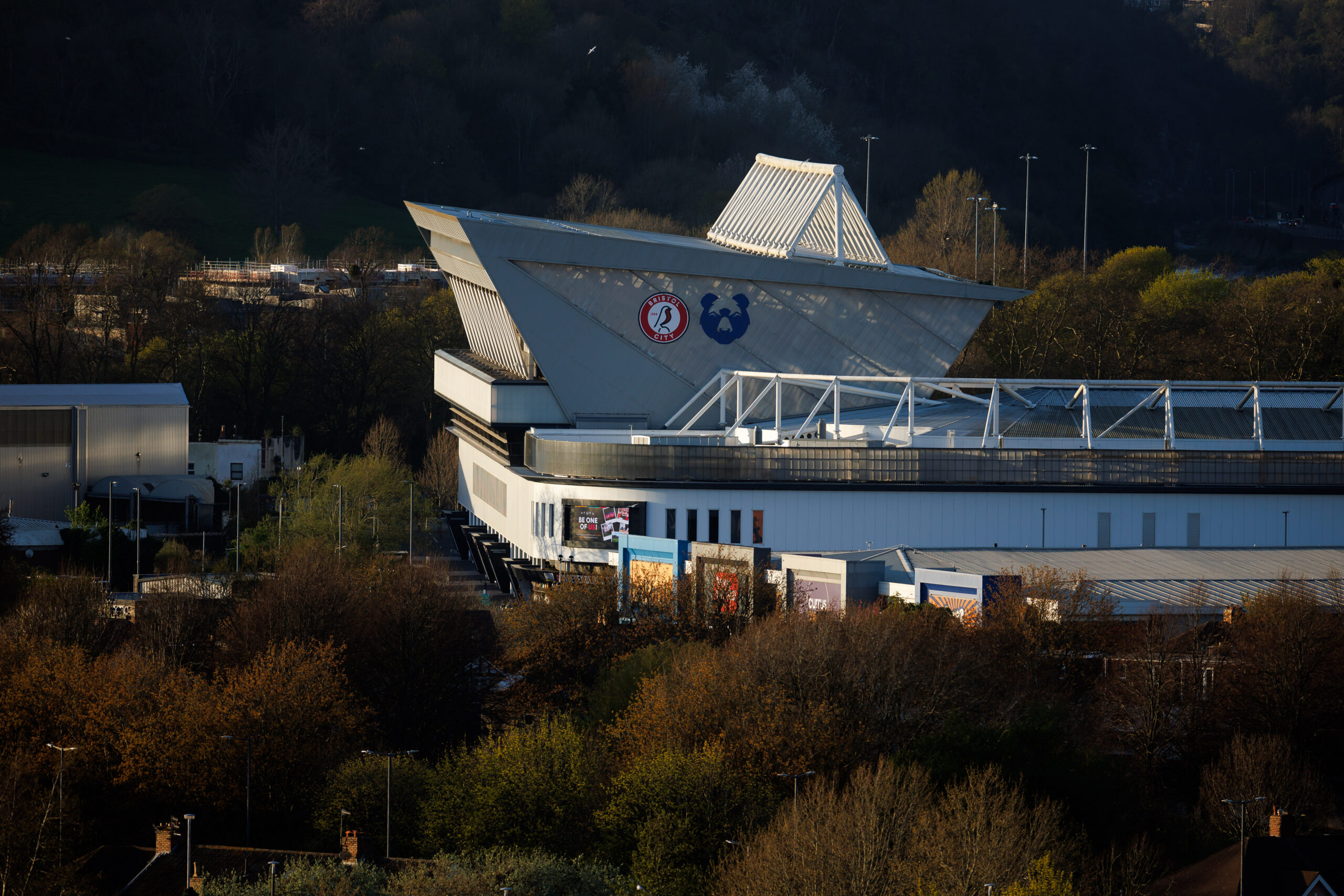 Modern sports stadium with a distinctive triangular roof and large logos on its facade, surrounded by trees and buildings under a clear sky.