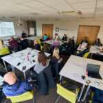 A group of adults sit at tables in a brightly lit conference room, attending a presentation projected onto a screen. Summer Modeshift Regional Networking Events