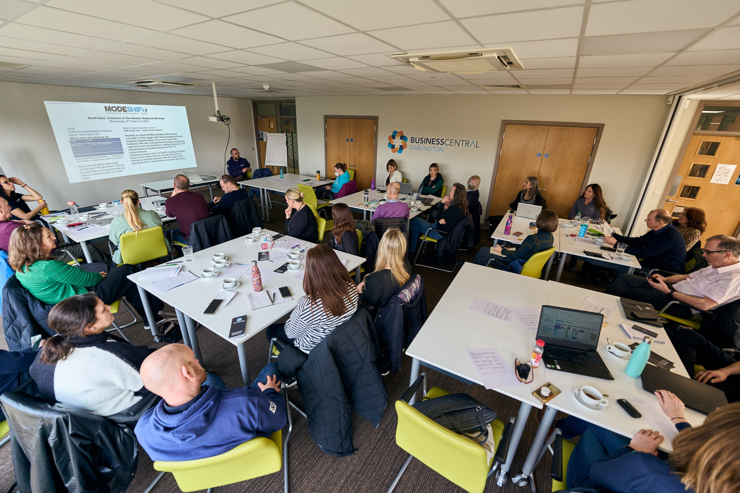 A group of adults sit at tables in a brightly lit conference room, attending a presentation projected onto a screen. Summer Modeshift Regional Networking Events