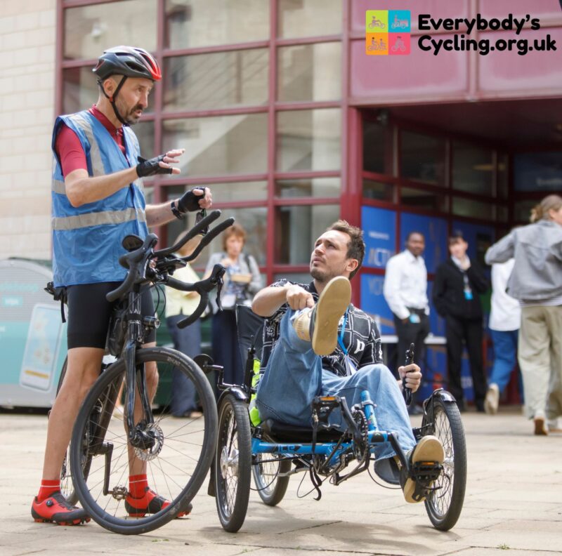 A man in a blue safety vest talks to another man seated in a blue recumbent tricycle, with a standard bicycle parked beside them.