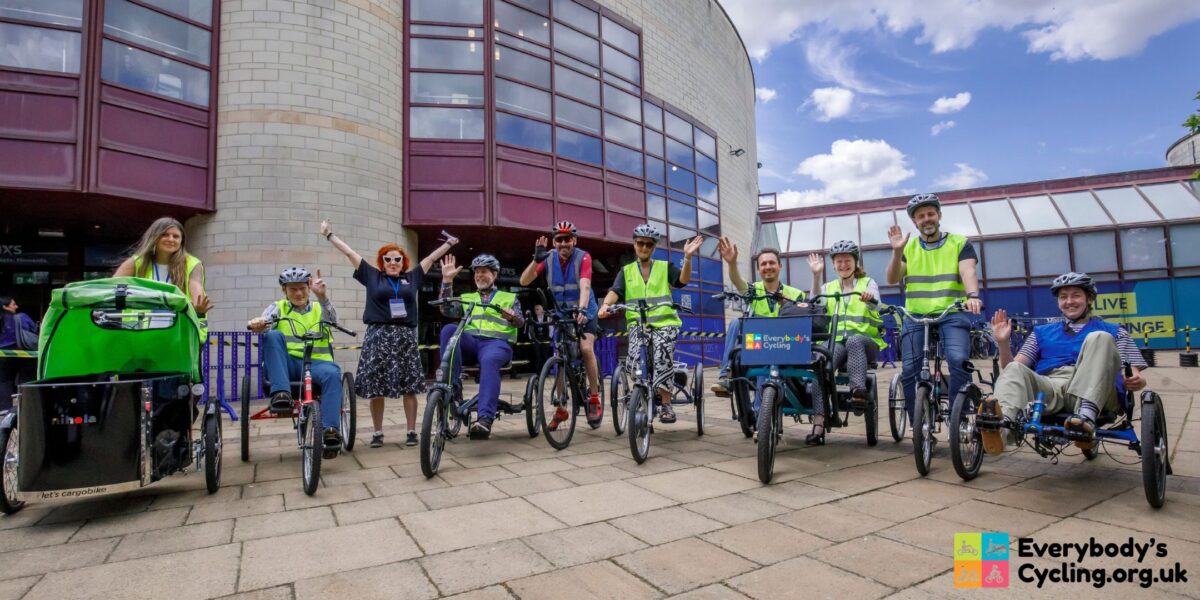 A group of people in high-visibility vests and helmets ride various types of bicycles and trikes outside a building with large windows, waving to the camera.