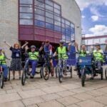 A group of people in high-visibility vests and helmets ride various types of bicycles and trikes outside a building with large windows, waving to the camera.