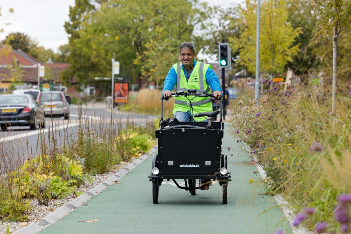 A person wearing a bright yellow safety vest rides a black electric cargo bike down a green bike path, with a busy road and trees in the background.