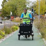 A person wearing a bright yellow safety vest rides a black electric cargo bike down a green bike path, with a busy road and trees in the background.