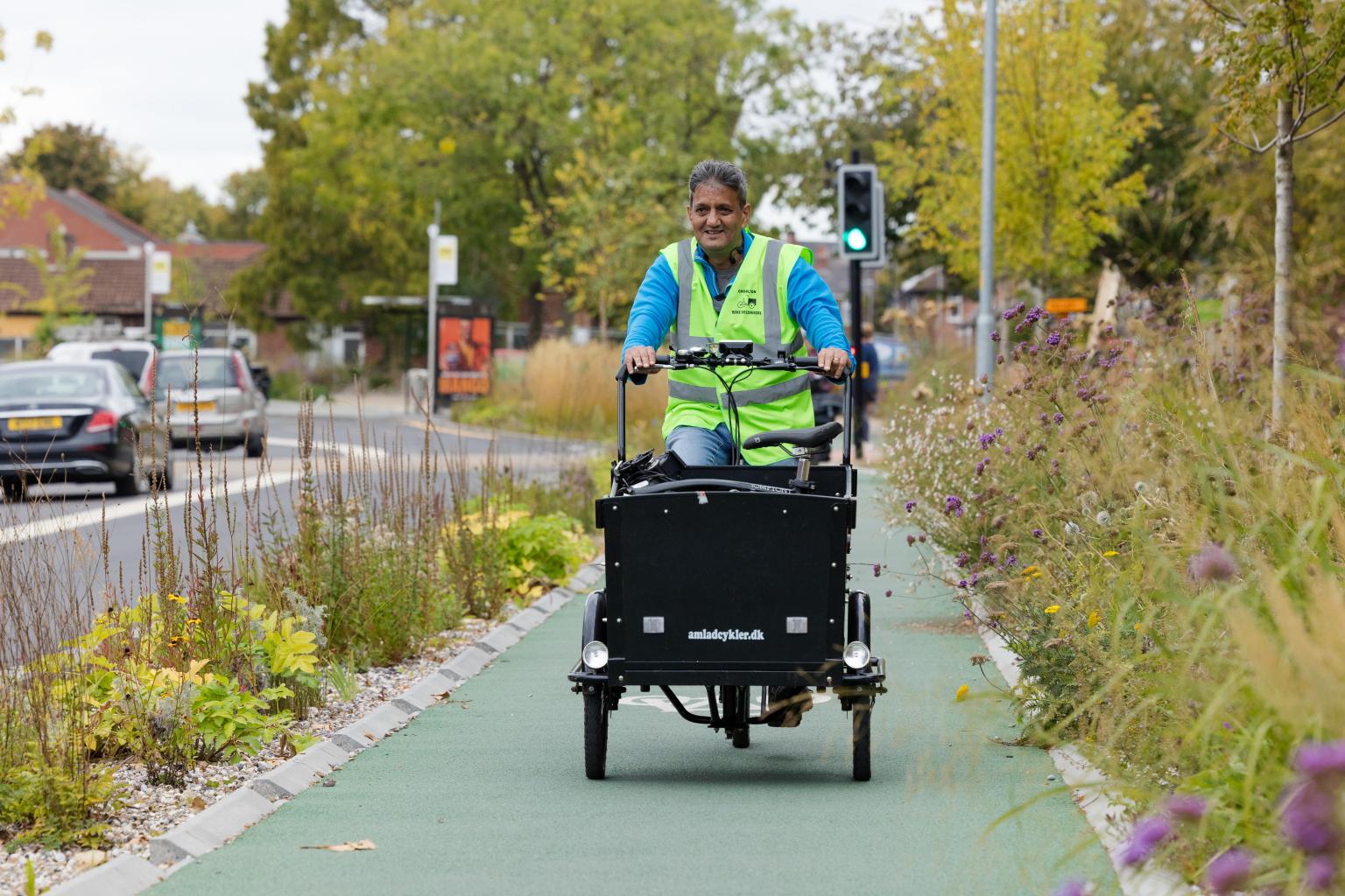A person wearing a bright yellow safety vest rides a black electric cargo bike down a green bike path, with a busy road and trees in the background.