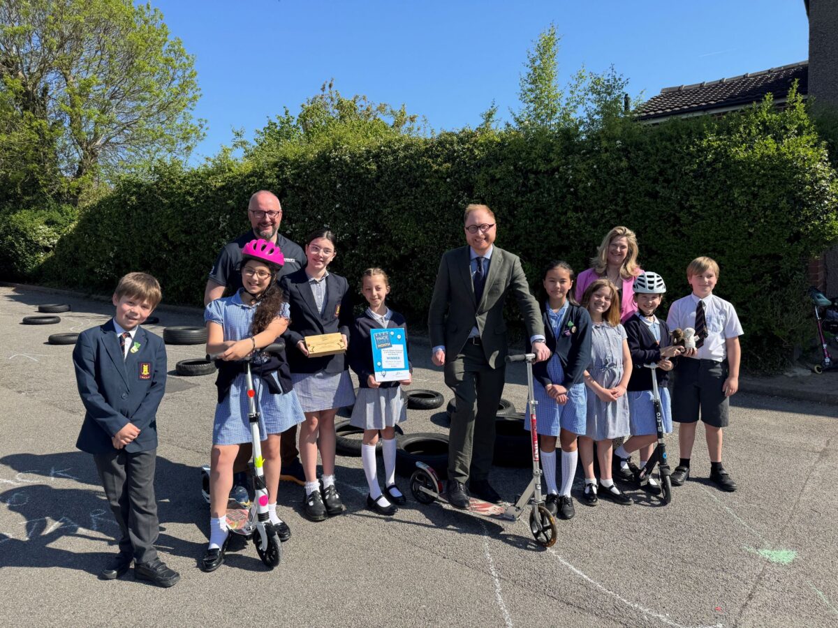A group of school children and adults pose for a photo with scooters and awards in an outdoor paved area with hedges and houses in the background.