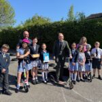 A group of school children and adults pose for a photo with scooters and awards in an outdoor paved area with hedges and houses in the background.