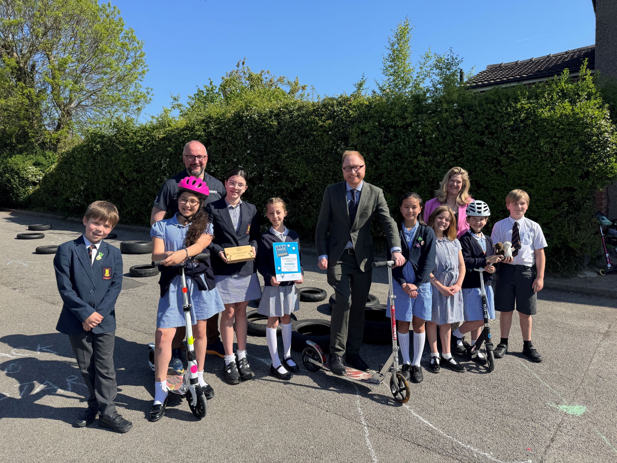 A group of school children and adults pose for a photo with scooters and awards in an outdoor paved area with hedges and houses in the background.