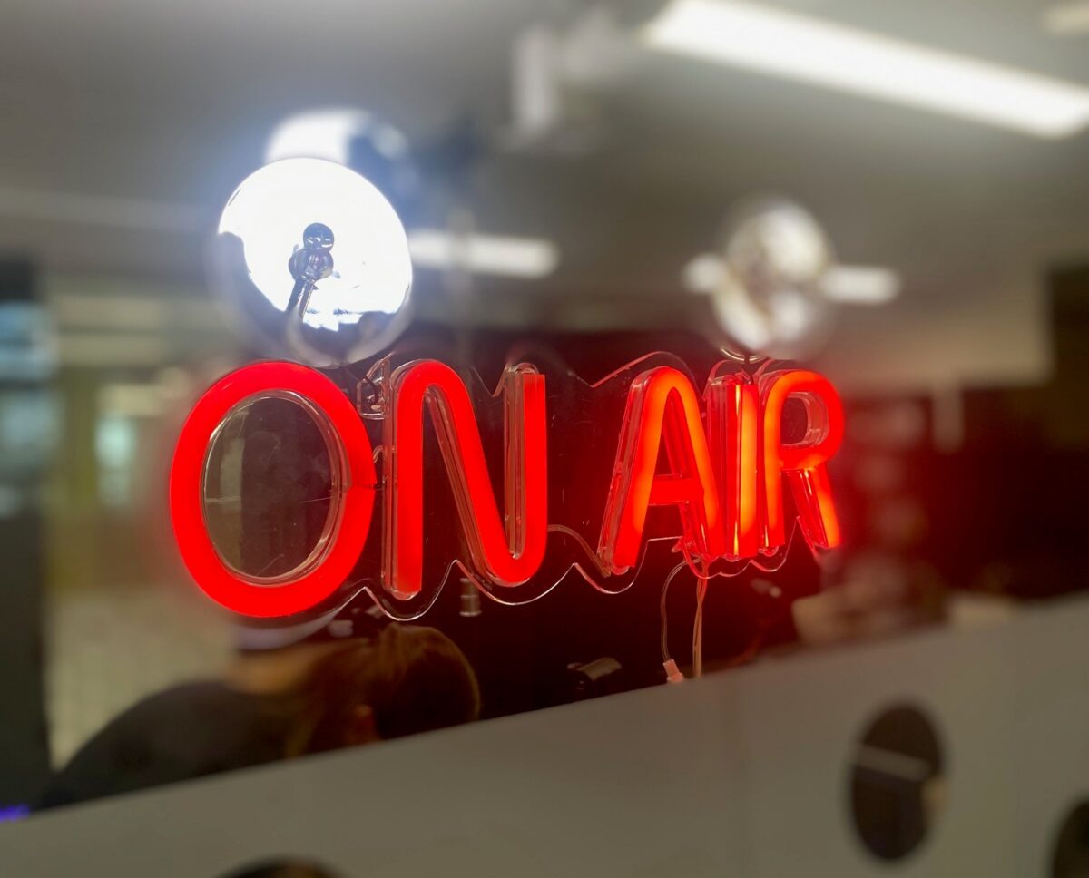 A neon sign with the text lit up in red - reads: 'On Air'. A studio in the background.