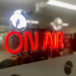 A neon sign with the text lit up in red - reads: 'On Air'. A studio in the background.