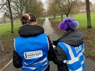 Two people wearing blue "Active Travel Inspectors" vests walk along a paved path in a park with trees on either side.