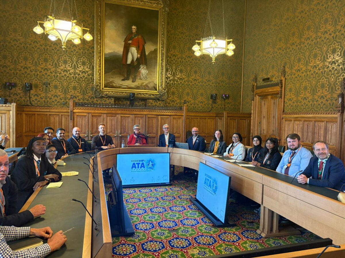 A group of people, including young adults are seated at a long, curved table in a room with ornate golden wallpaper and a large portrait of a man in uniform. Two large screens displaying "MODSHIFT ATA" are visible on the table.