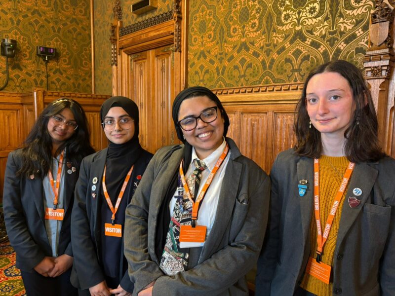 Four young people wear suits and lanyards with "VISITOR" badges, standing in a room with ornate wood paneling and patterned wallpaper.