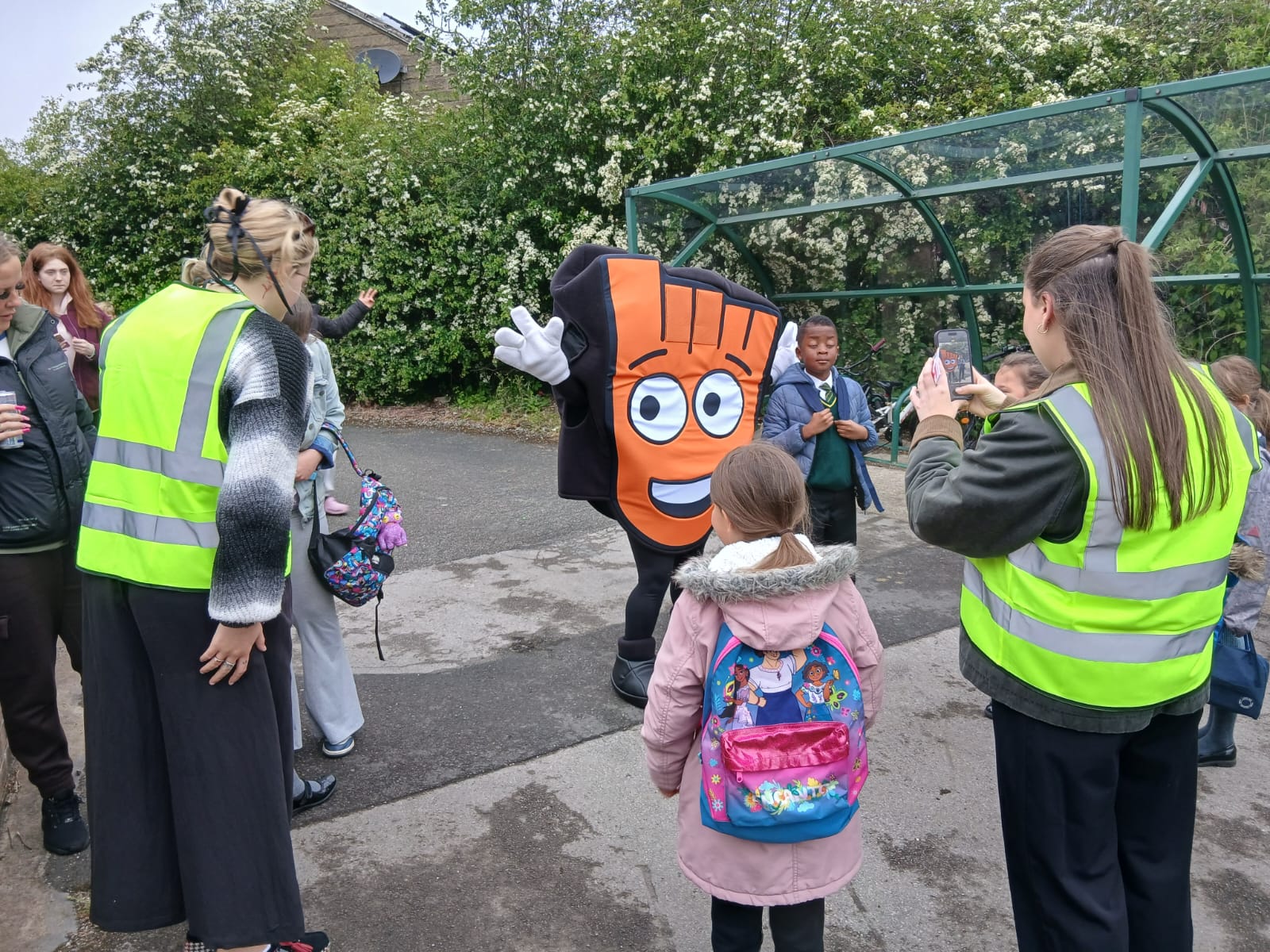 A mascot in an orange and black costume with a large smiling face waves to a group of people, some wearing high-visibility vests, outdoors near a covered cycle parking space.