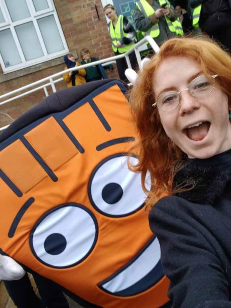 A young woman with red hair takes a selfie with an orange cartoon character costume.