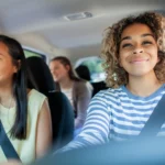 Three young women smiling and wearing seatbelts inside a car, one in the driver's seat.